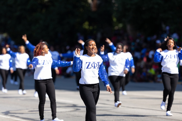 A group of Zeta Phi Beta members walk down the street in a parade. They wear matching blue and white Zeta Phi Beta crewnecks.