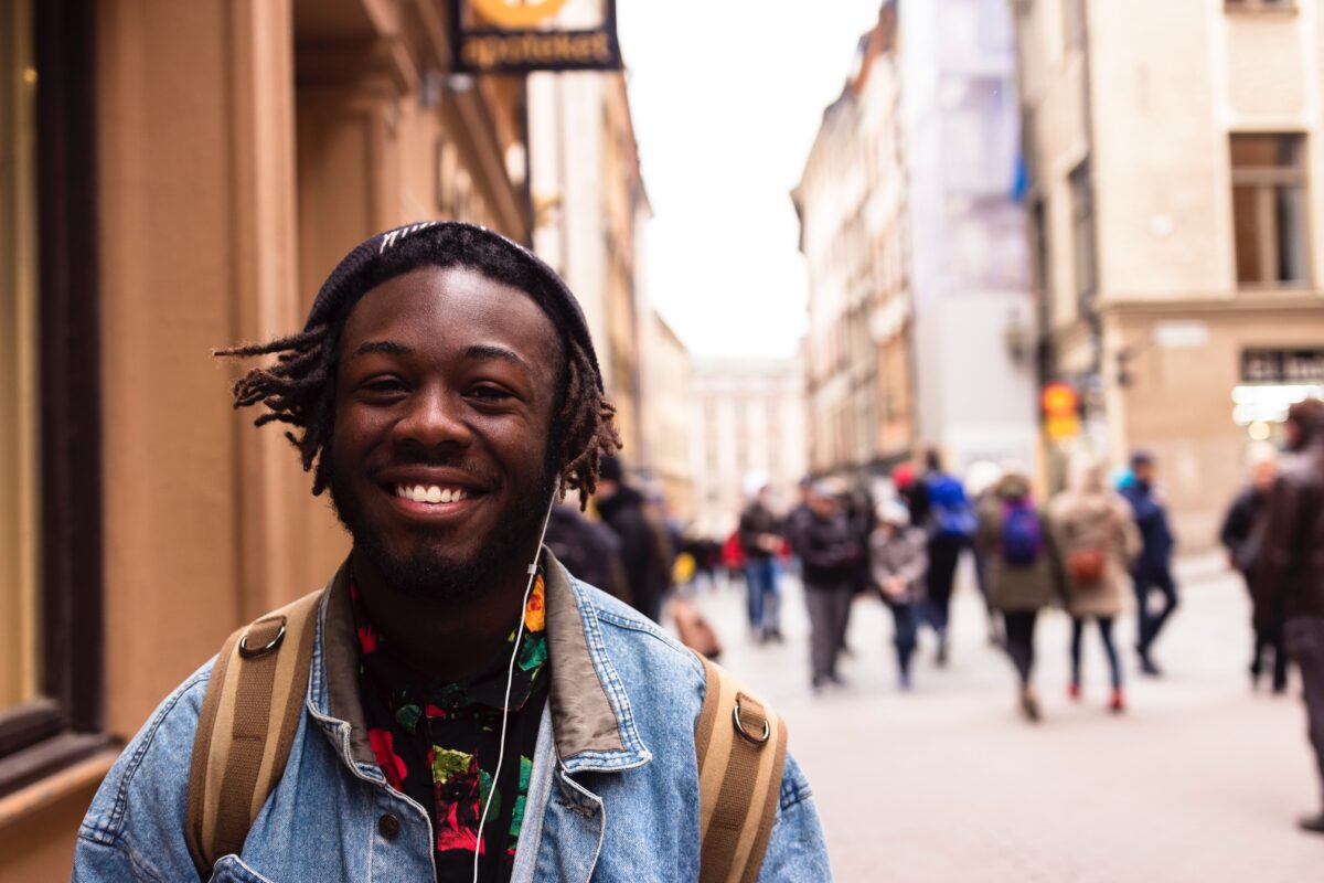 A young man with brown skin and black locs stands on the street smiling at the camera. The background is blurry but people walk up and down the street. He wears a backpack, jean jacket, and beanie.