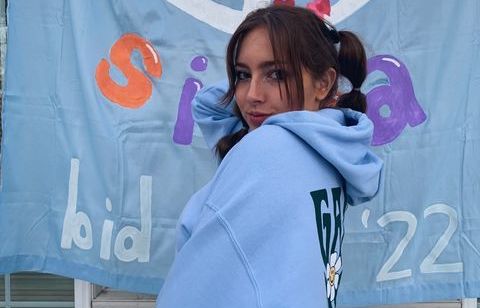Maddie (a young woman with light skin and brown hair) poses in front of a blue painted banner for Gamma Delta Sigma bid day.