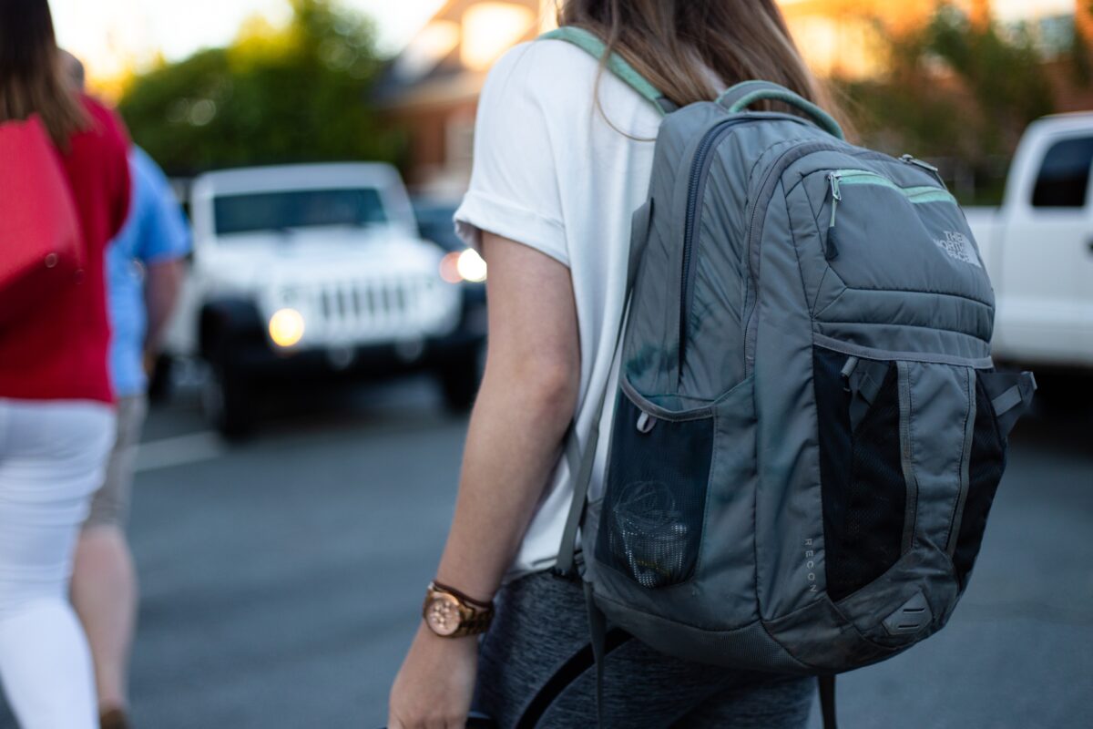A girl with light skin and brown hair walks down the street wearing a backpack.