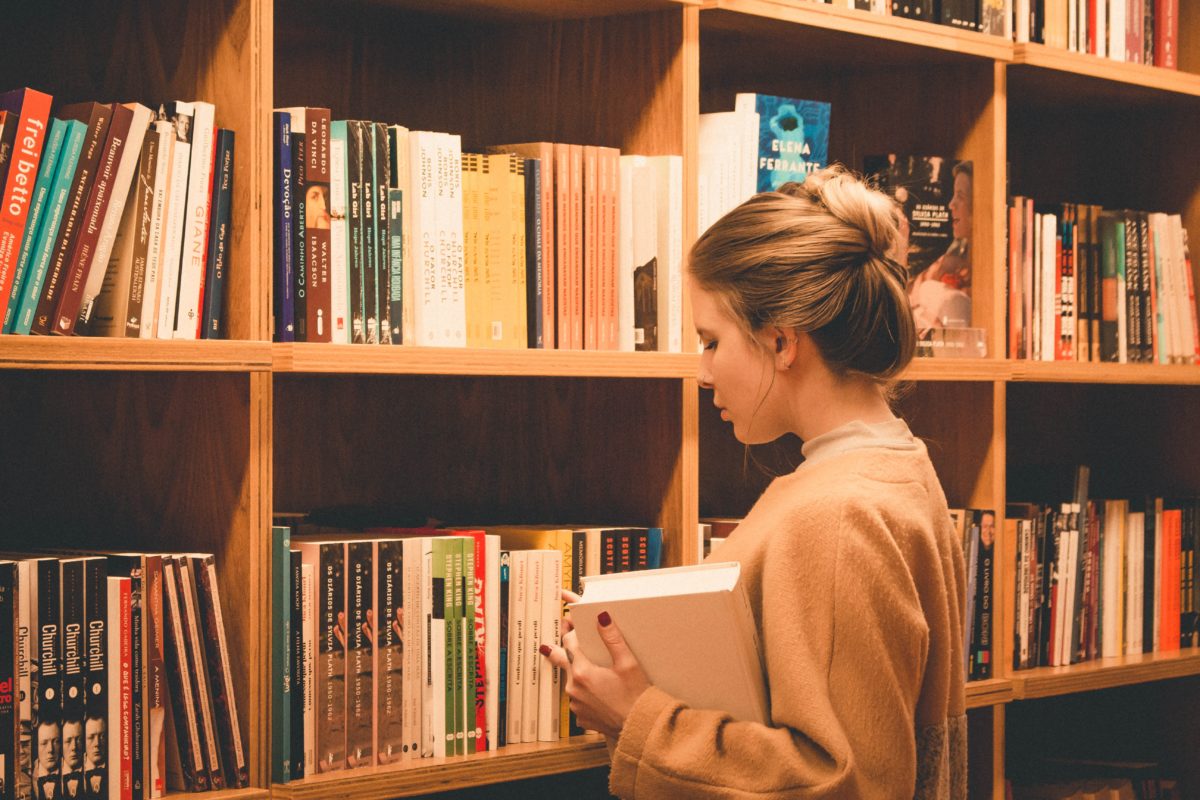A young woman with light skin and blond hair pulls a book out of a bookshelf.