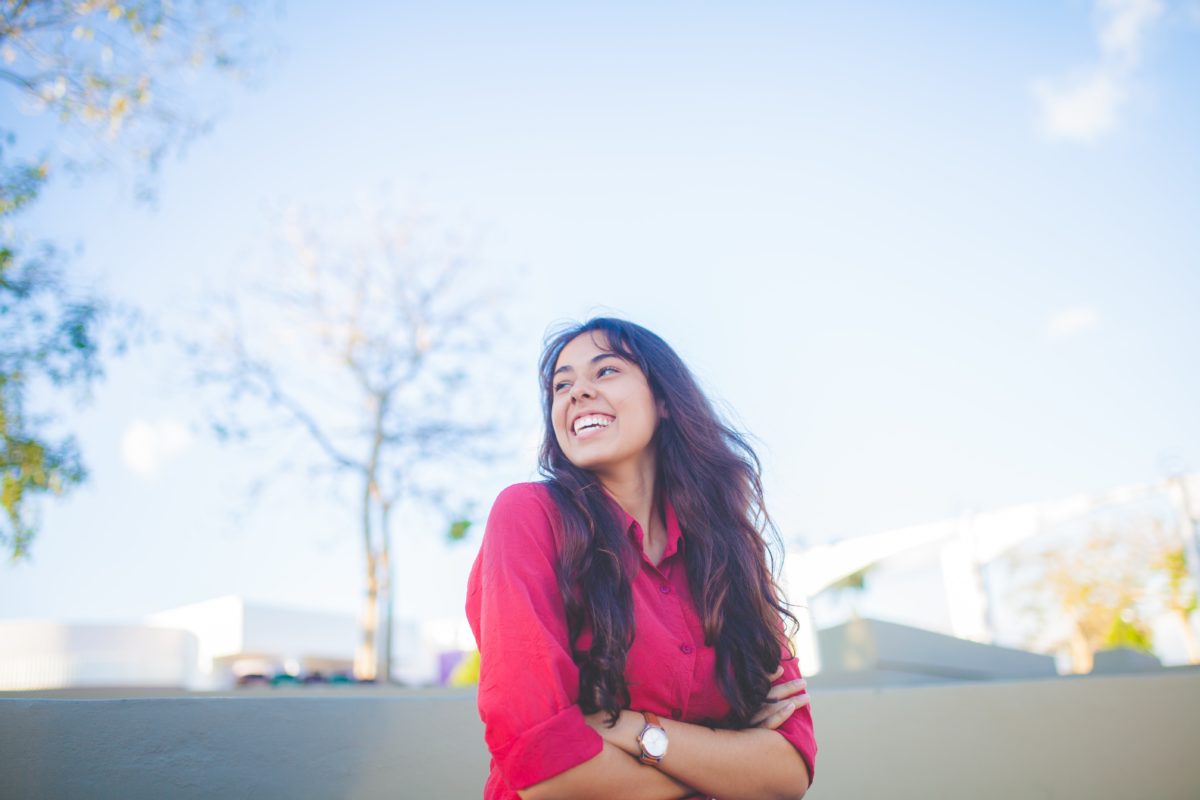 A young woman with brown skin and hair crosses her arms and smiles. She stands outside in front of a clear sky. 