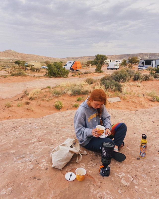 A young woman sitting on the ground of a desert eating. Campers are parked behind her.