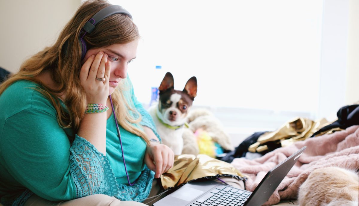 A young woman sits on her bed with her laptop in her lap. Behind her is a dog looking at the camera.