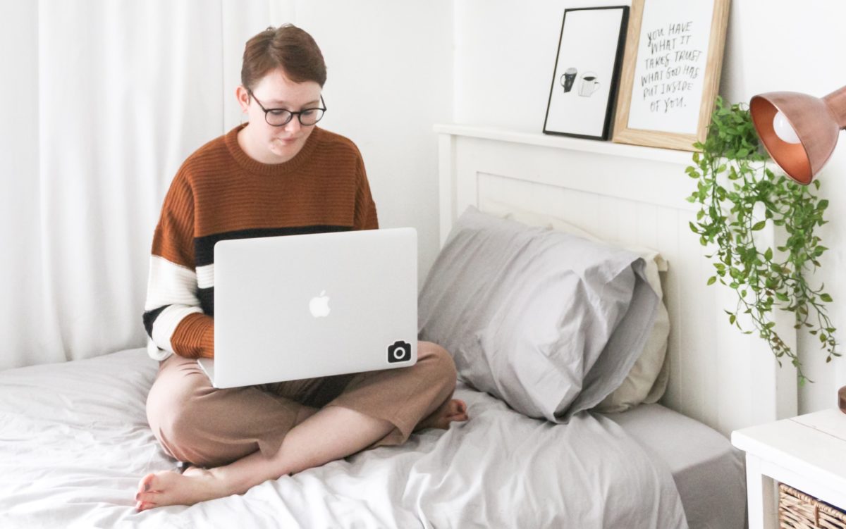 A young woman sits crosslegged on her bed looking at her laptop.