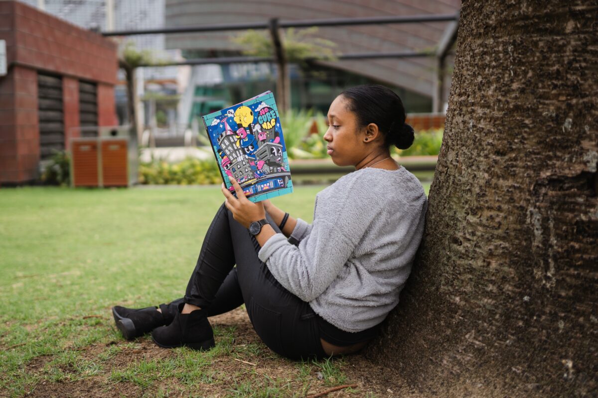 A young woman leans on a tree reading a colorful book.