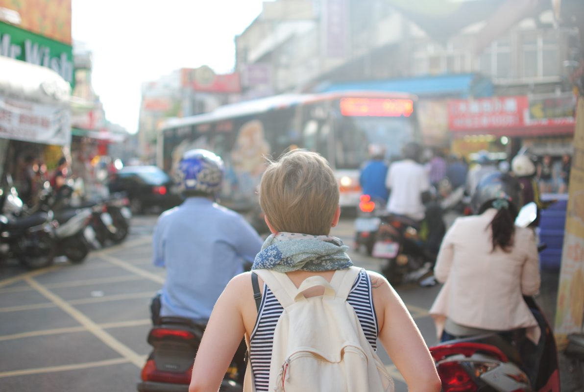 A young woman stands facing a street filled with buses and mopeds.