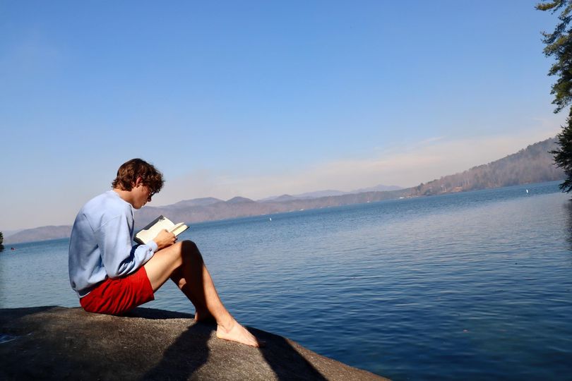 A young man sits on a rock reading a book on a lake shore.