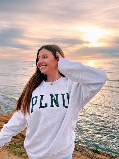 A young woman poses in a PLNU sweatshirt in front of an ocean view.