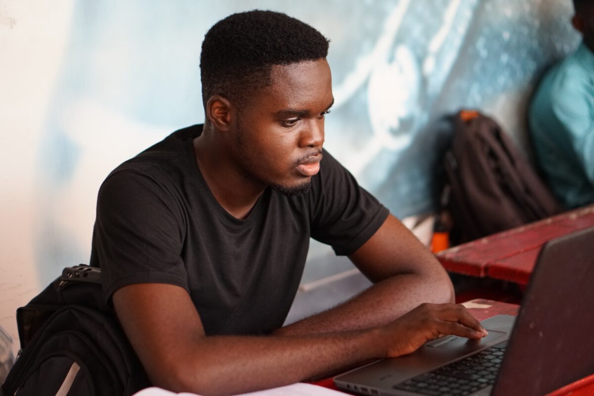 A young black man sits at a table working on a laptop.