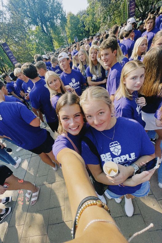 Two young women take a selfie in a crowd of students wearing matching purple t-shirts.
