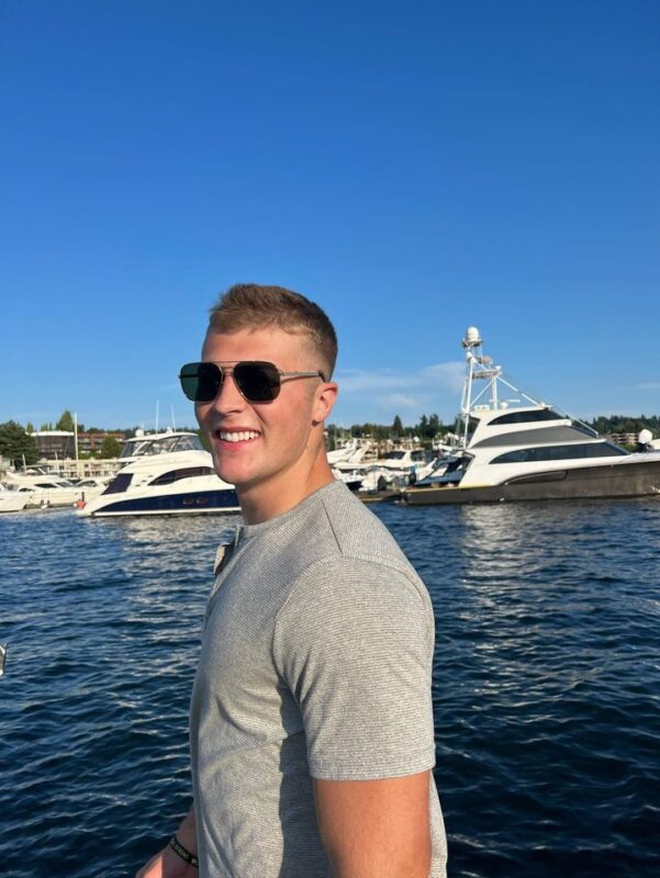 A young man poses in front of a port filled with boats.