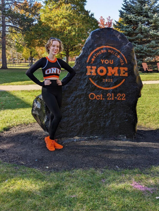 A young woman in Ohio Northern University merch stands in front of a boulder painted to say "Northern Welcomes You Home"